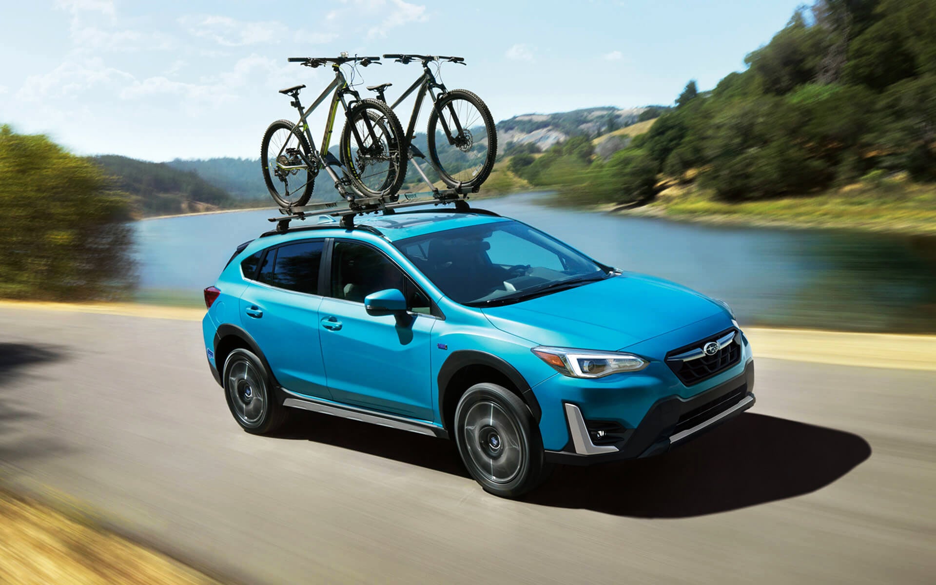 A blue Crosstrek Hybrid with two bicycles on its roof rack driving beside a river | Valley Subaru of Longmont in Longmont CO